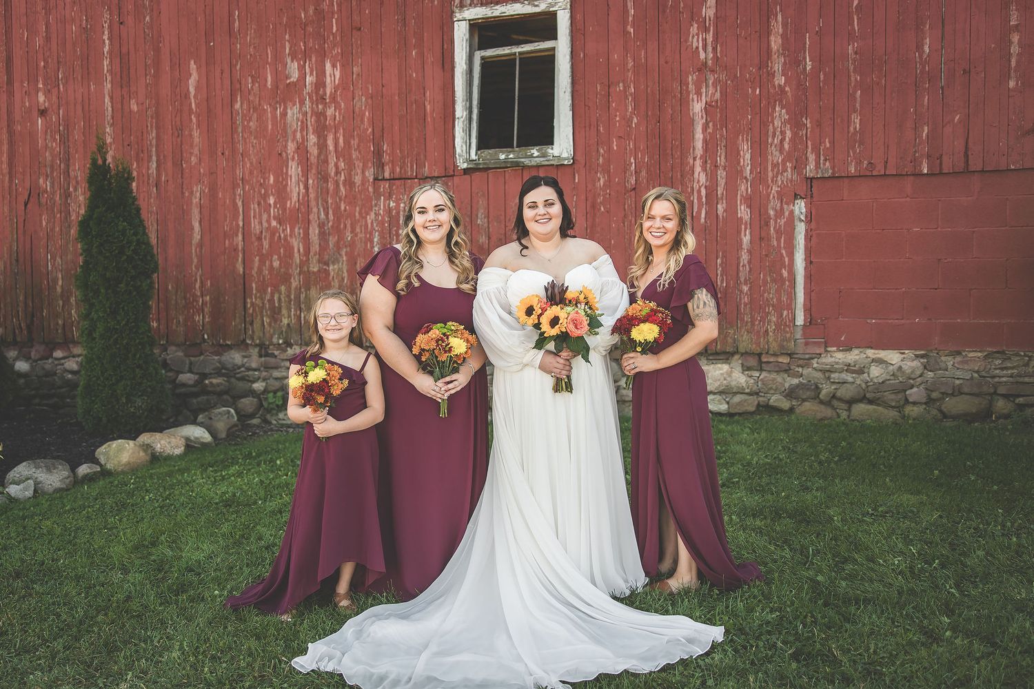 Bridal party in burgundy dresses poses with bride in white against red barn.