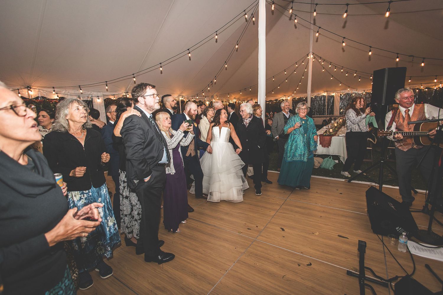 Large wedding tent with string lights and wooden dance floor filled with celebrating guests.