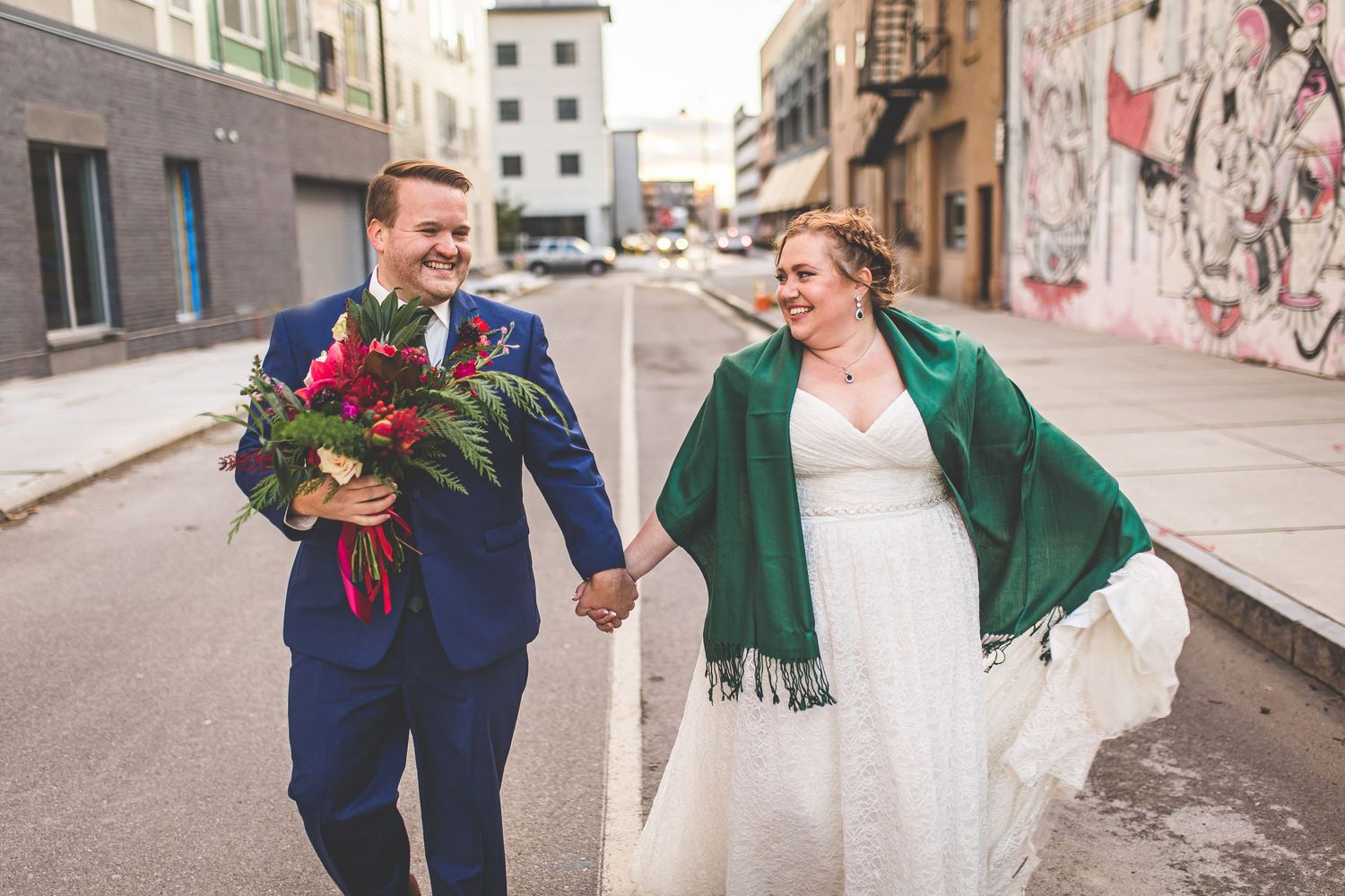 Newlyweds walking together on urban street with graffiti wall, holding red floral bouquet.