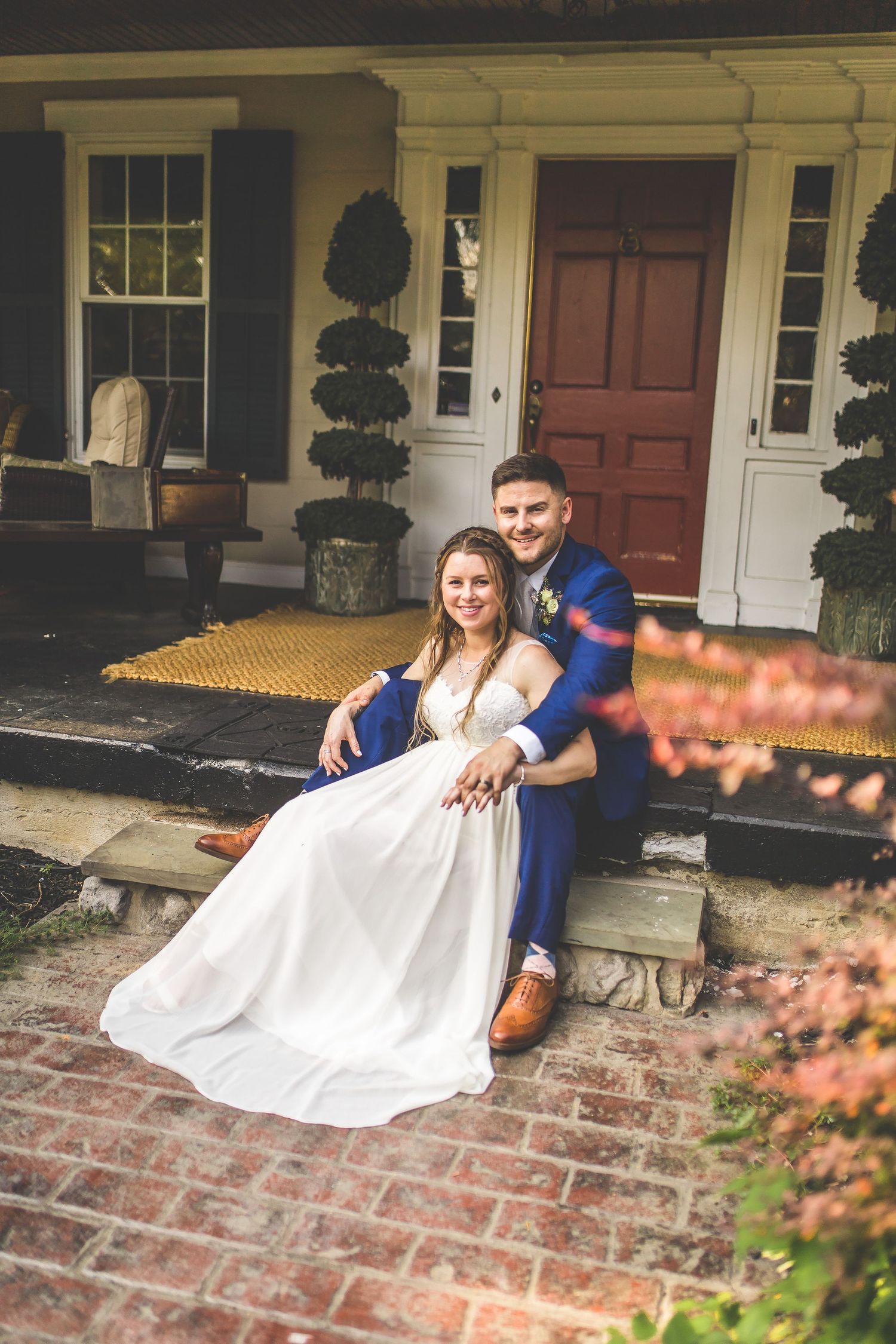 A couple in wedding attire sitting together on brick steps of a historic house with topiary decorations.