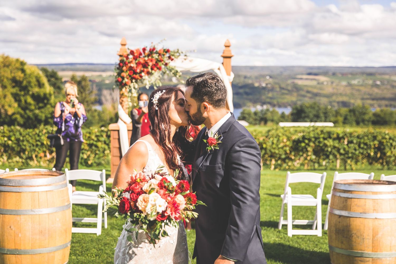 Wedding ceremony setup at a vineyard with wine barrels and white chairs under cloudy skies.