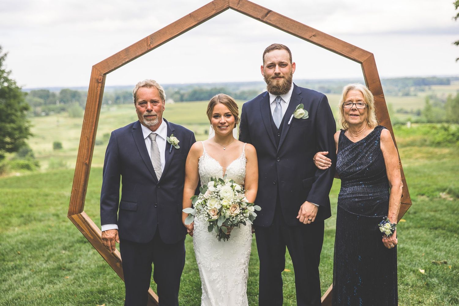 Family wedding portrait under wooden hexagonal arch with scenic countryside background.