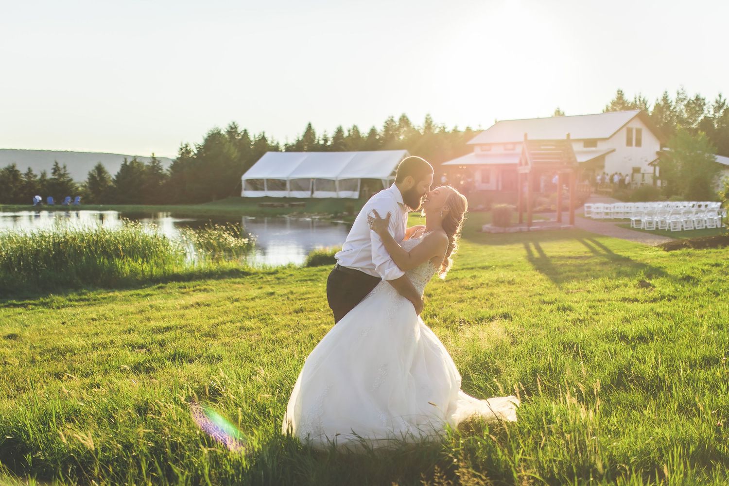 Sunset wedding portrait of couple embracing in a field with farmhouse in background.