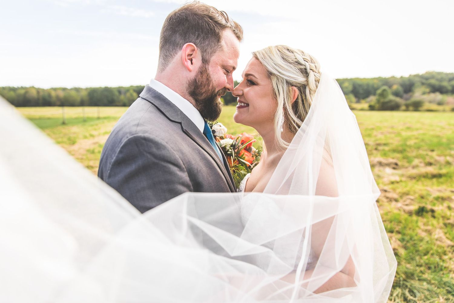 Romantic wedding portrait in sunlit field with flowing veil catching the breeze.