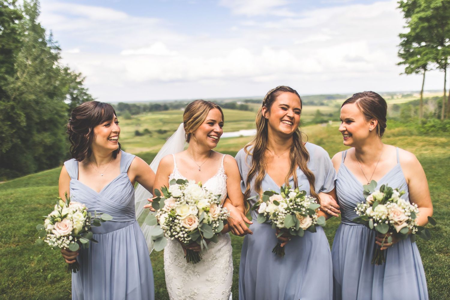 Bridesmaids in powder blue dresses laugh together while holding white floral bouquets.