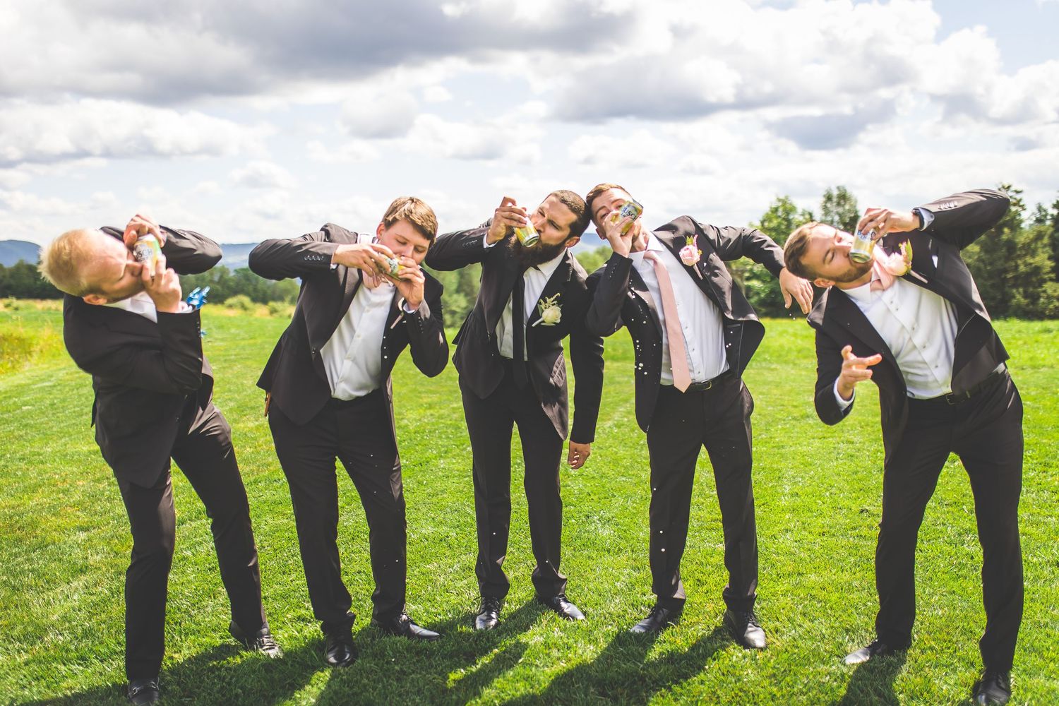 Groomsmen in black suits perform playful bending pose for group photo on grass.