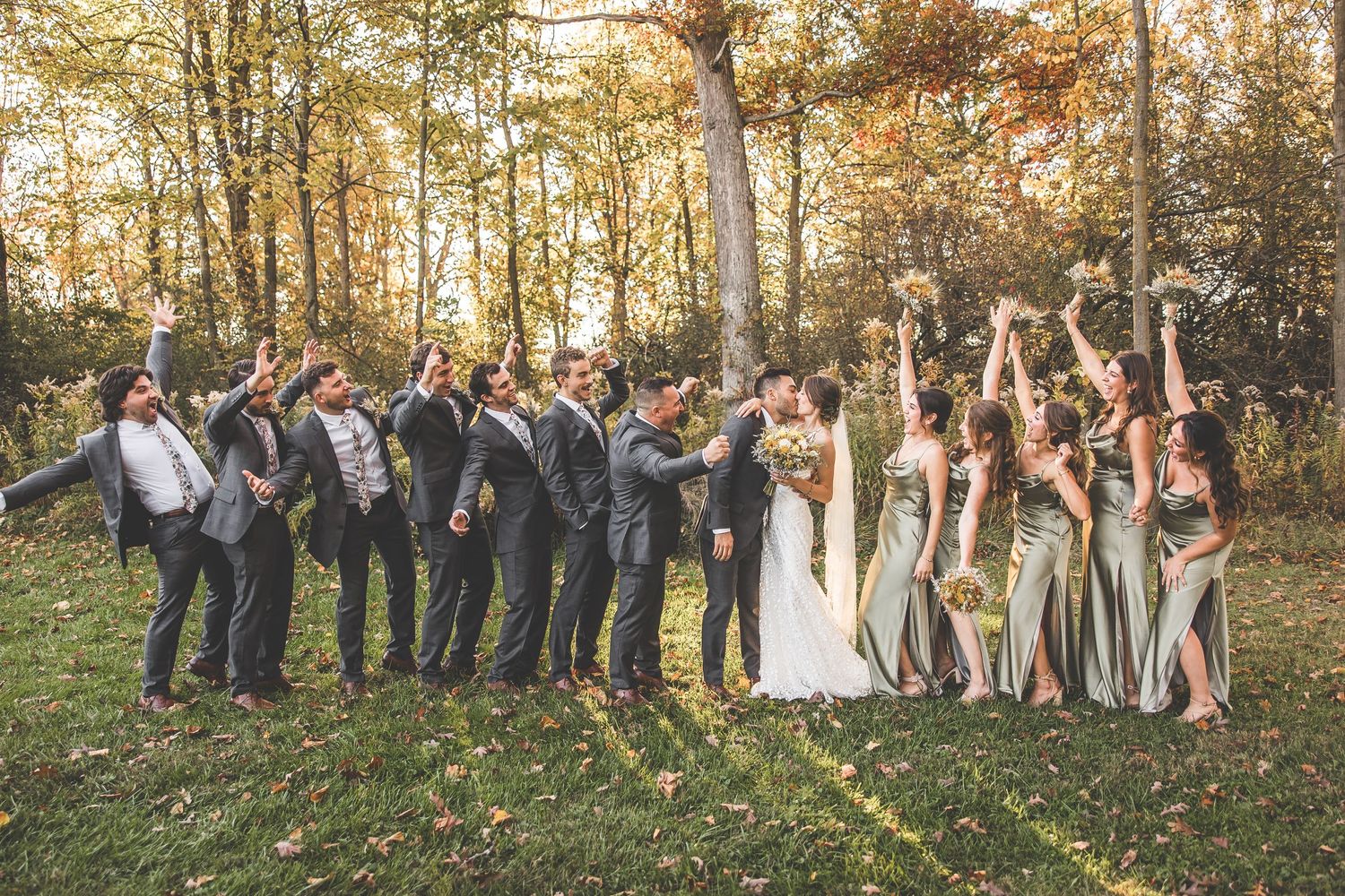Wedding party stands in line against backdrop of fall foliage and sunlit trees.