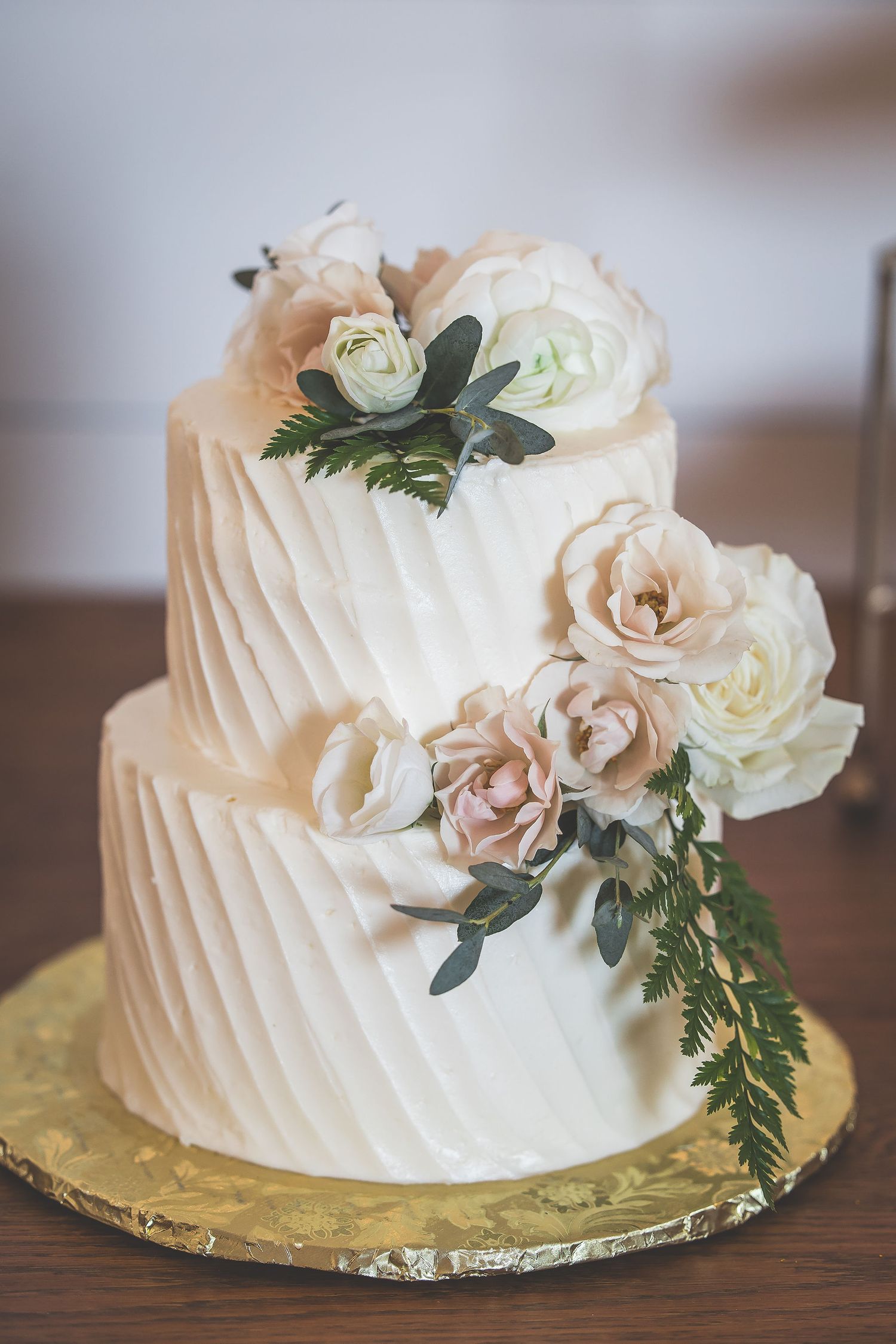Elegant white wedding cake decorated with blush roses and greenery on a gold cake board.