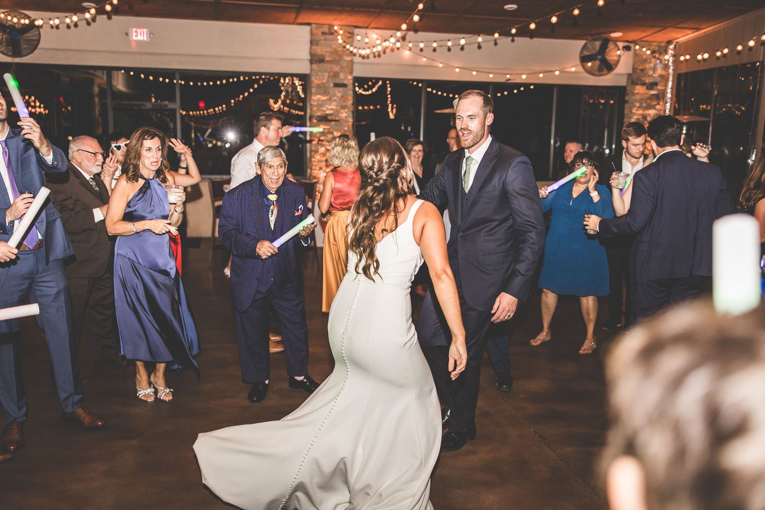 Guests celebrate on the dance floor at an indoor wedding reception with twinkling lights overhead.