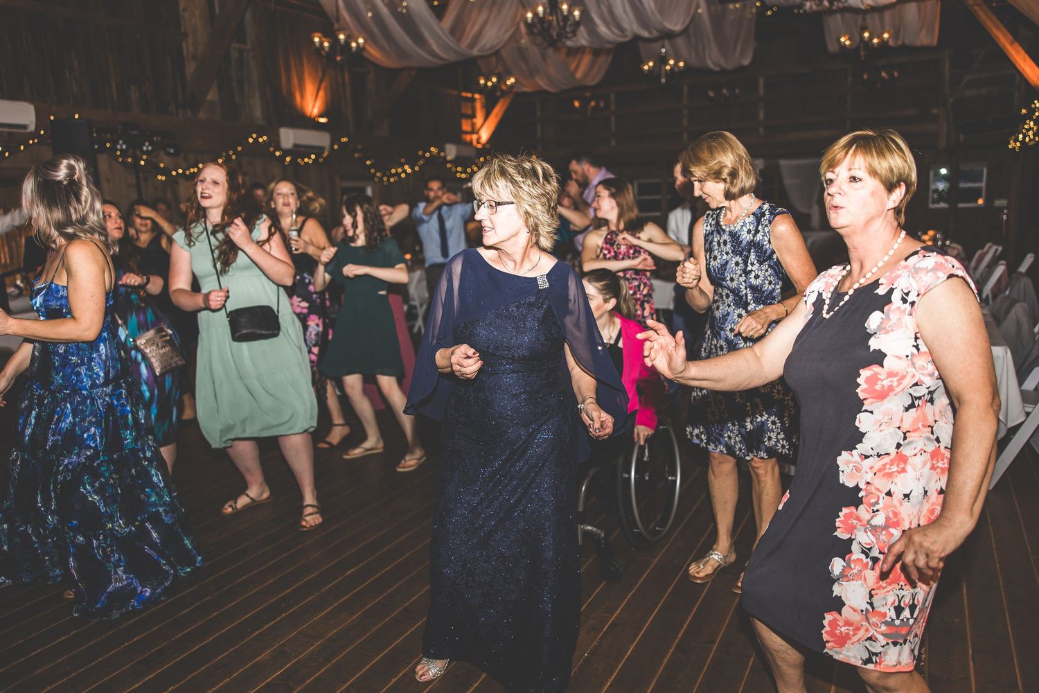 Wedding guests dance together on a wooden dance floor under warm lighting at an indoor reception venue.