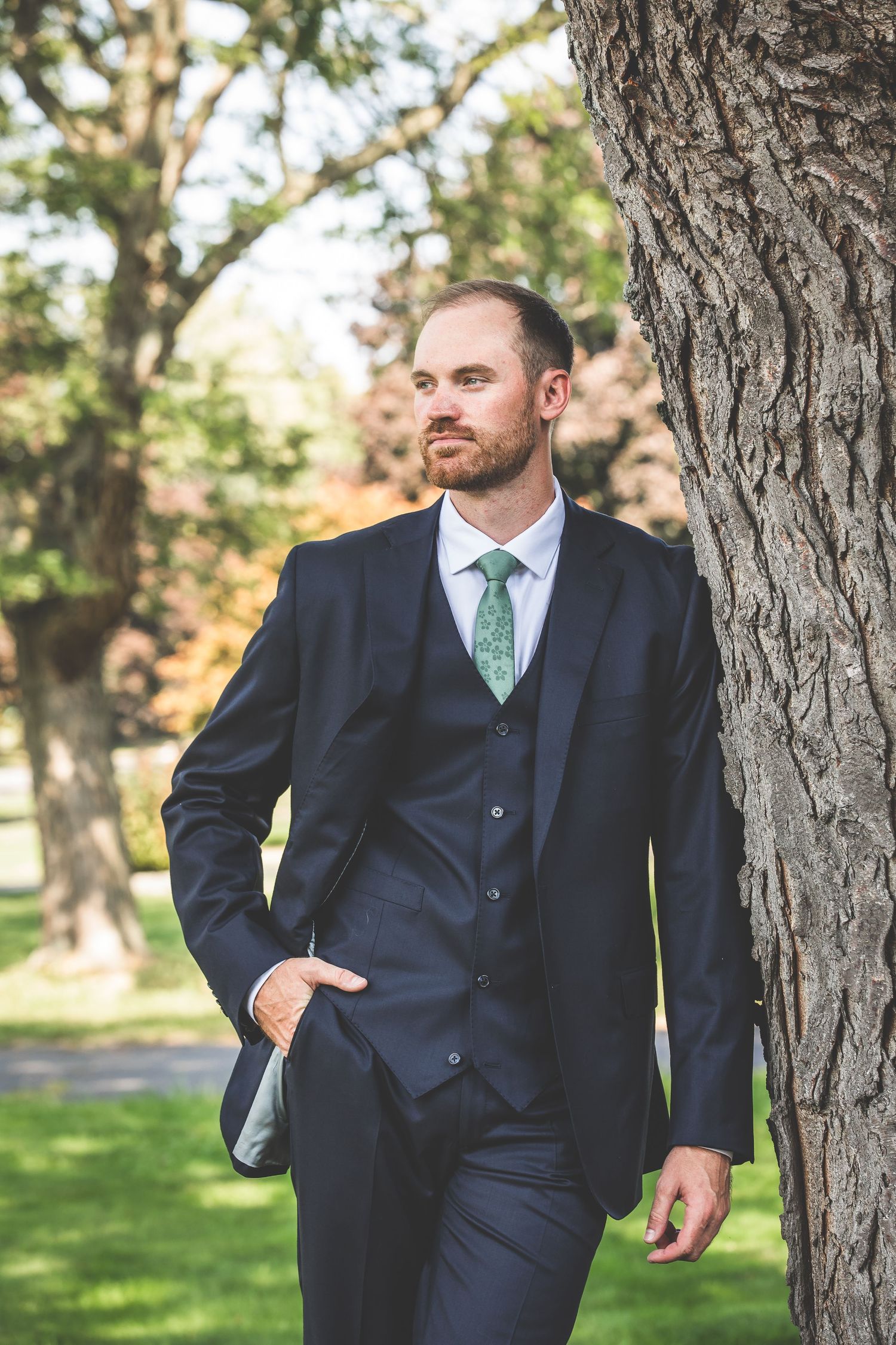 Groomsmen in dark suits and green ties stand casually between trees in an outdoor setting.