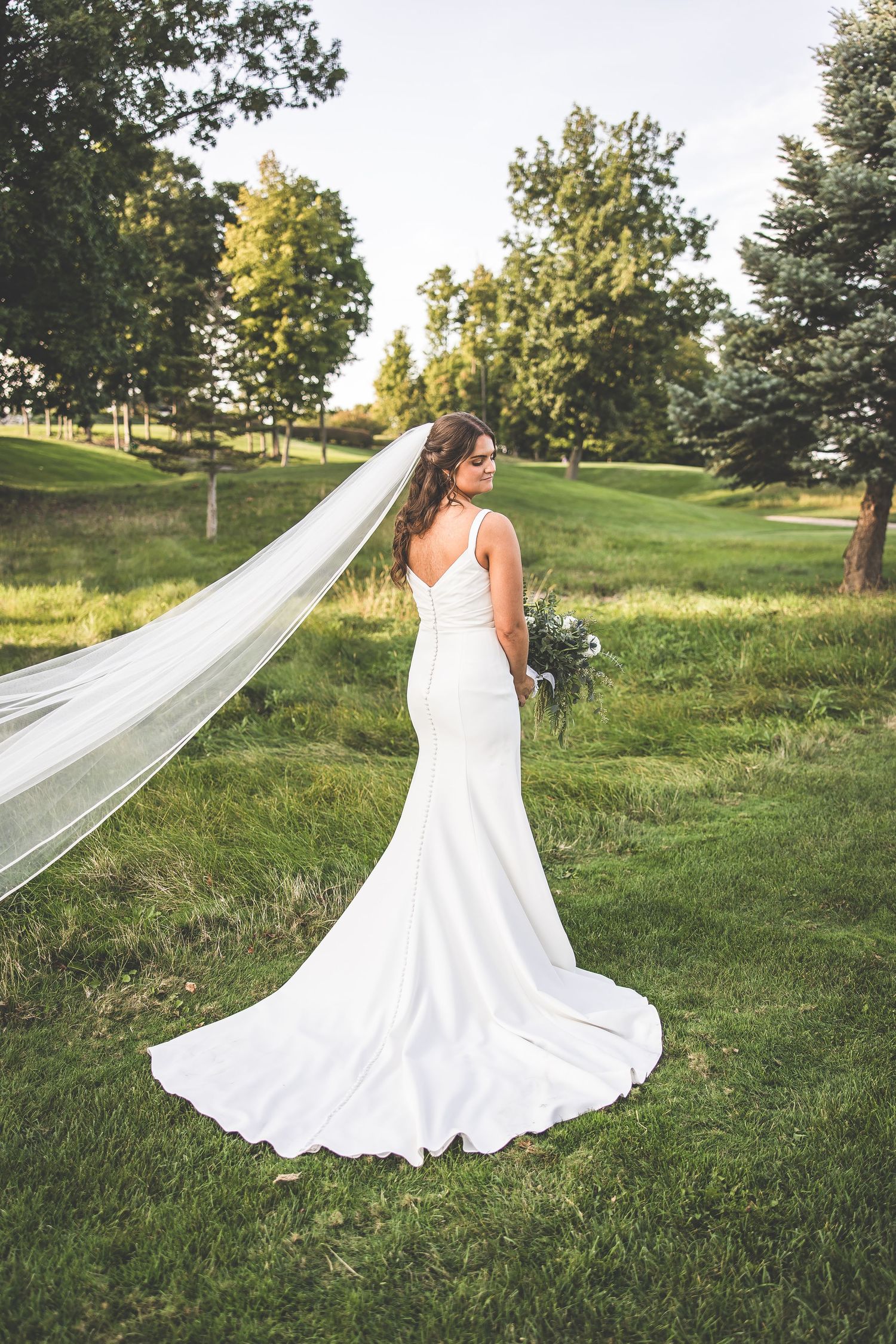 Elegant bride in fitted white gown with long veil stands in scenic outdoor setting.
