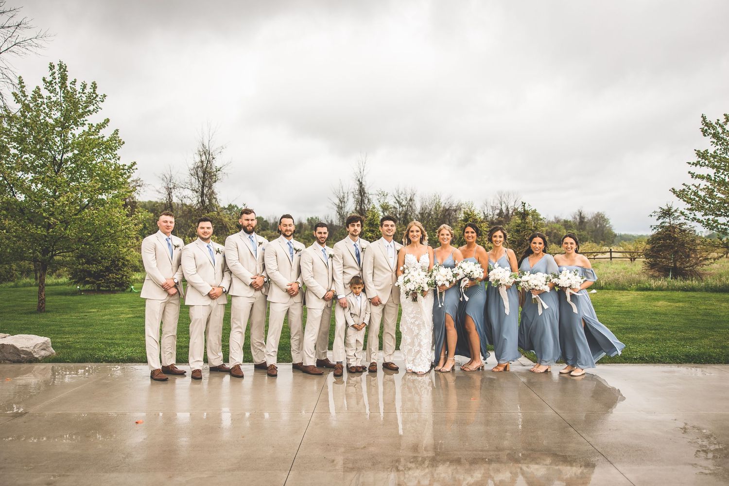Large wedding party in light beige and blue attire poses outdoors on rainy day.