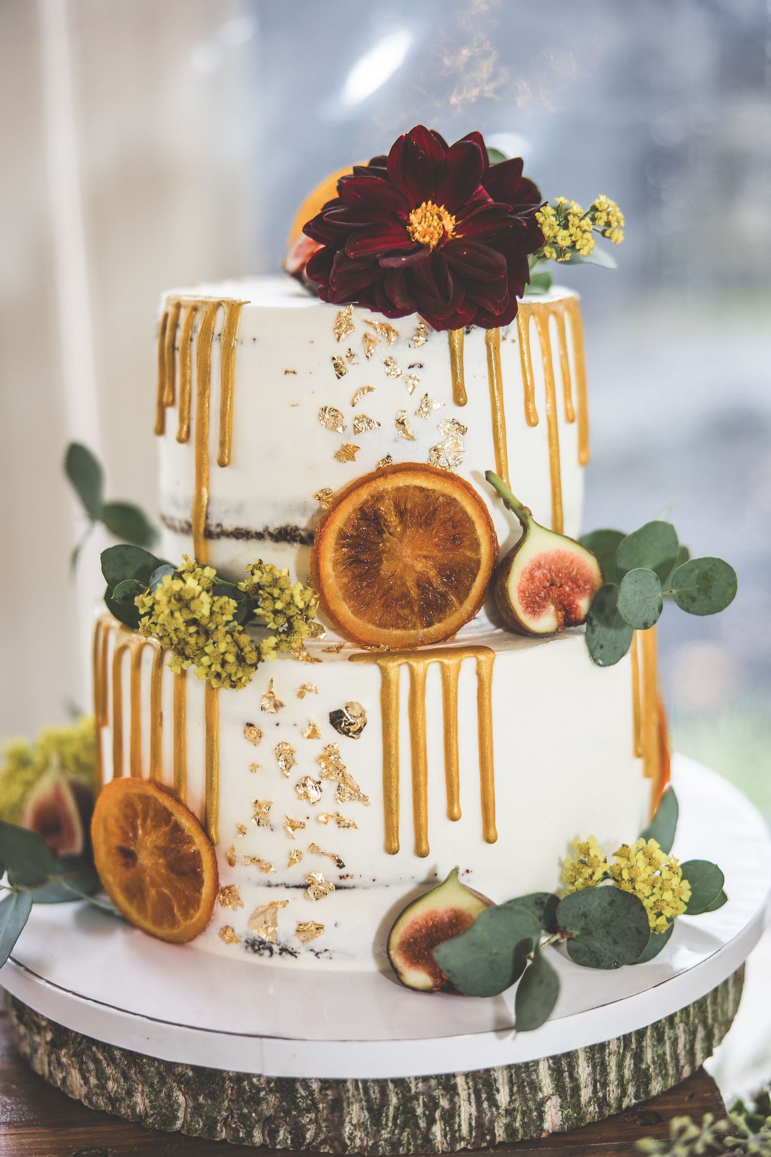 White wedding cake with gold drips decorated with dried orange slices, figs and burgundy flowers.