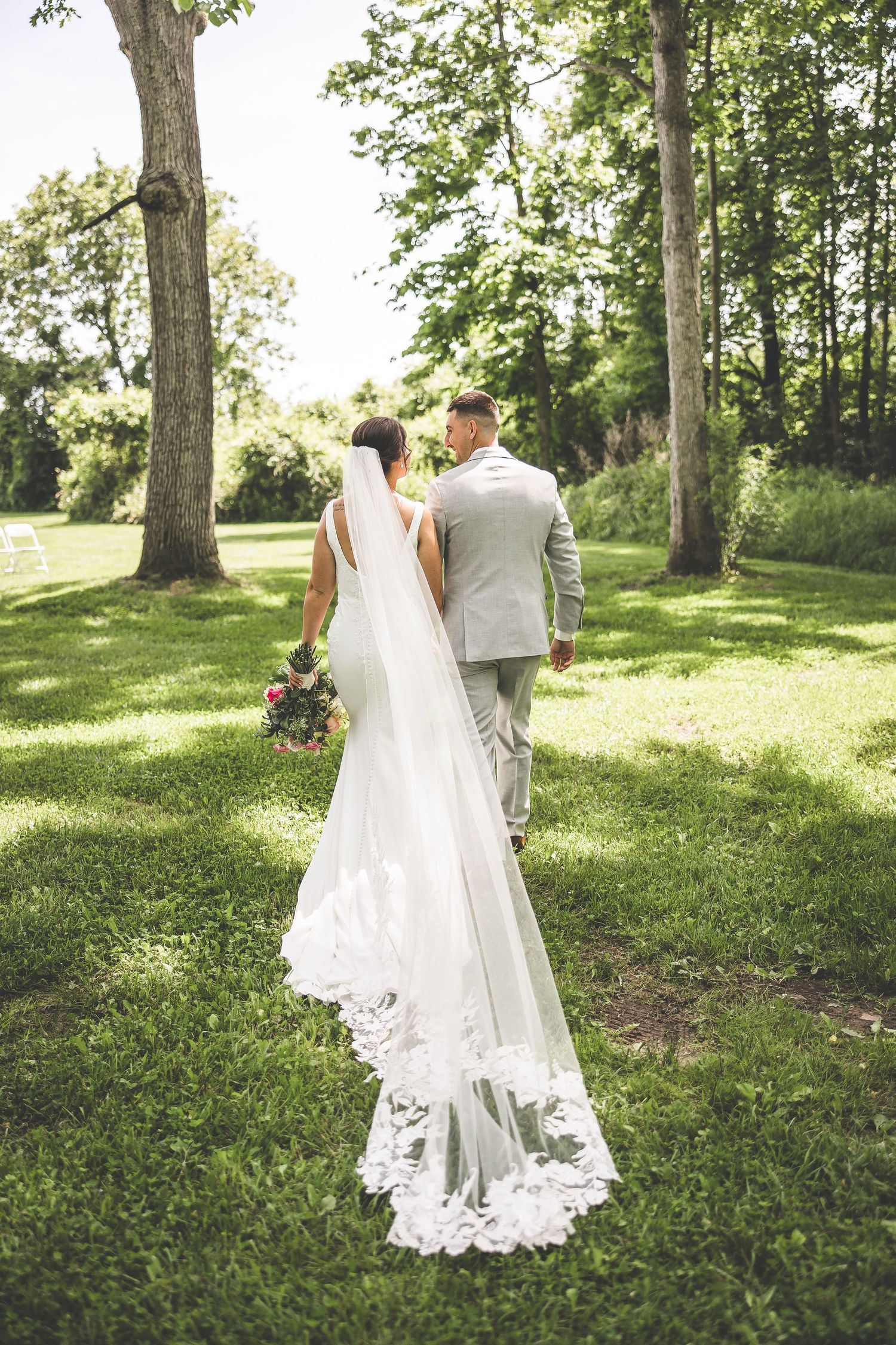 A long lace wedding veil trails behind a couple walking through a sun-dappled forest.