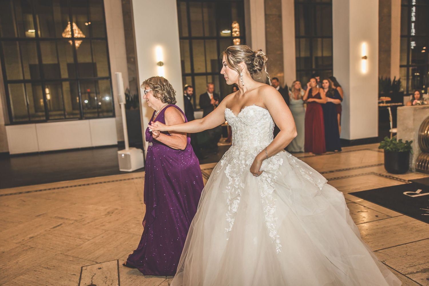 Bride in white lace gown dances with guest in purple dress during wedding reception.
