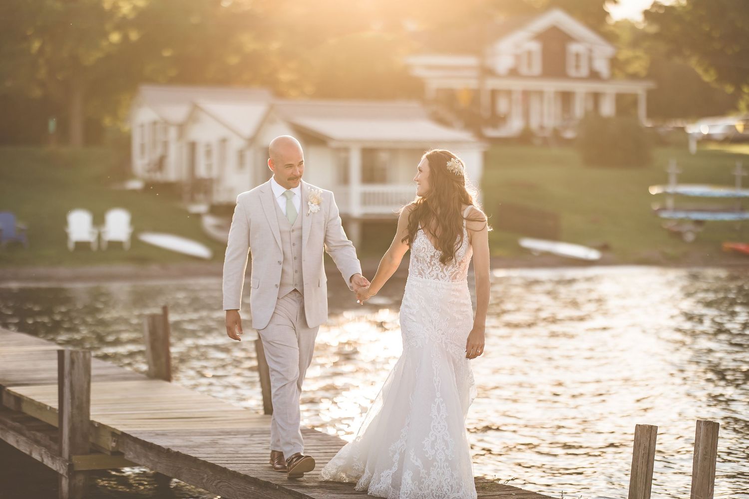 A couple walks hand in hand on a lakeside dock at sunset with houses in the background.