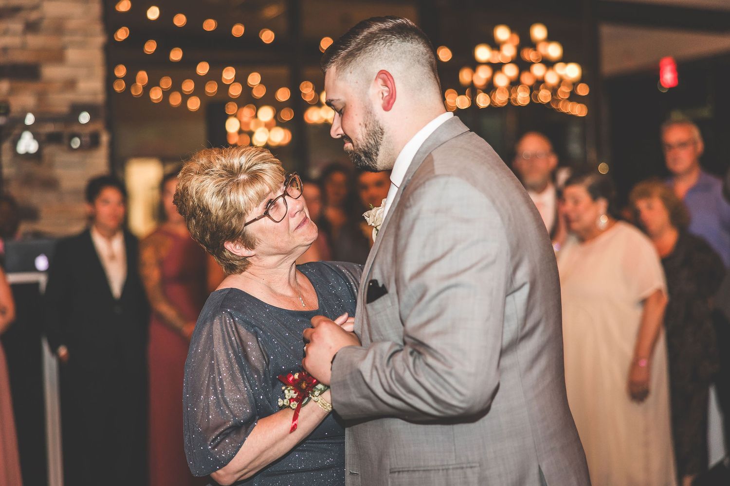 Wedding guests dance together under warm string lights at an evening reception celebration.
