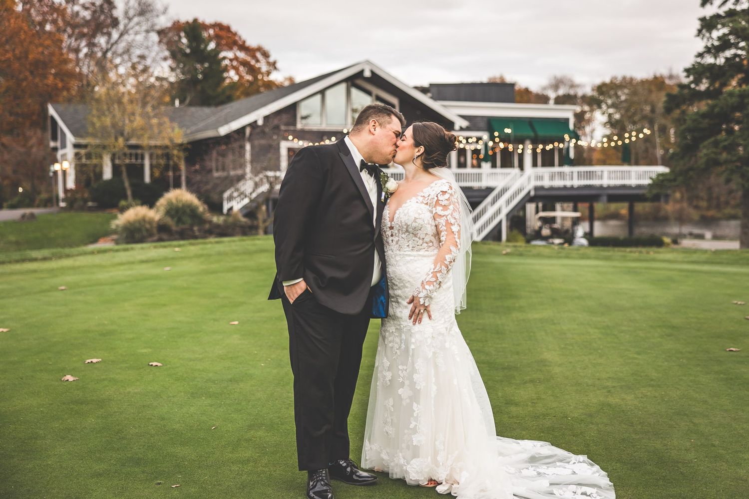 Wedding couple poses in front of a residential backdrop with fall scenery.