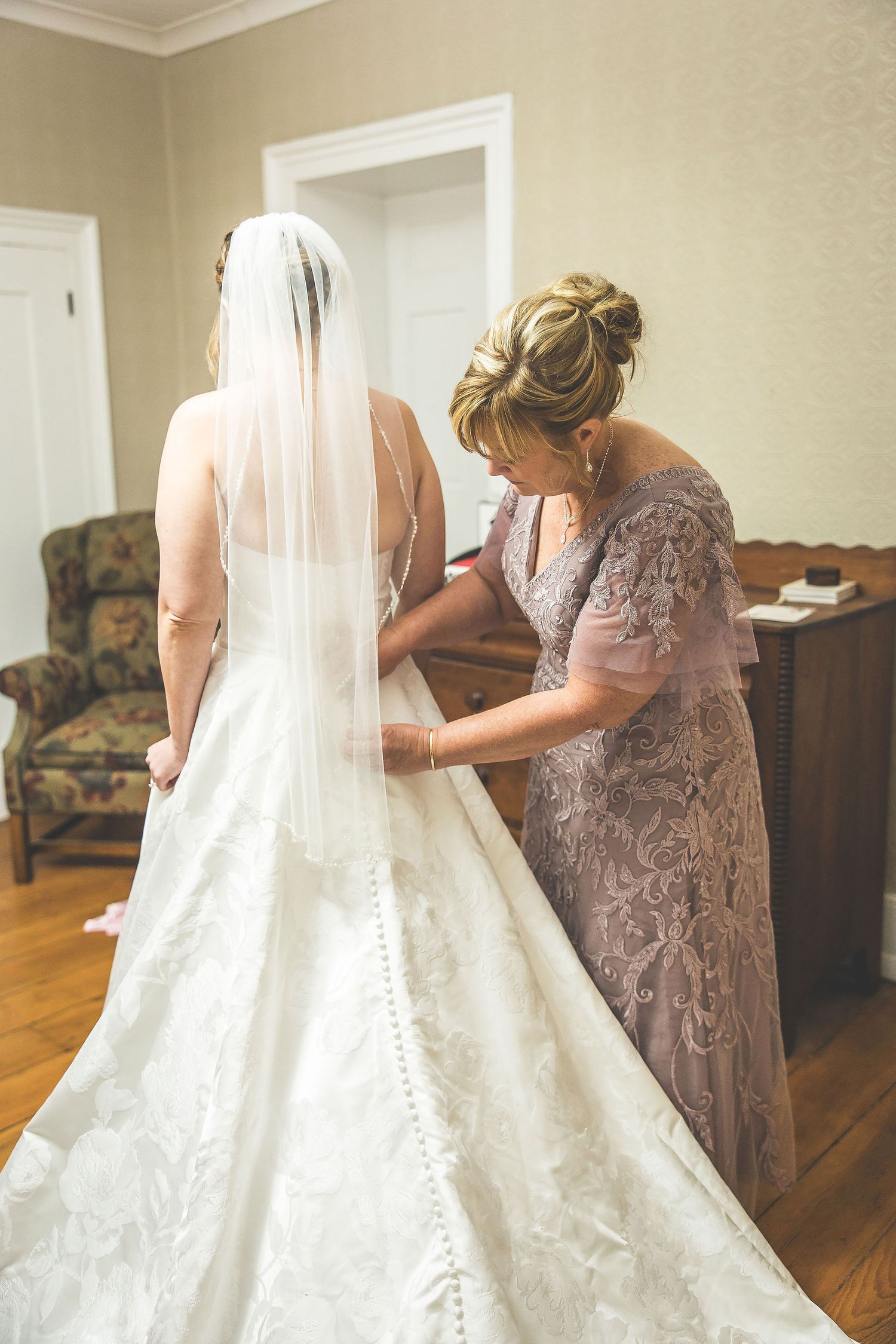 Wedding dress being adjusted in preparation room with hardwood floors and classic furniture.