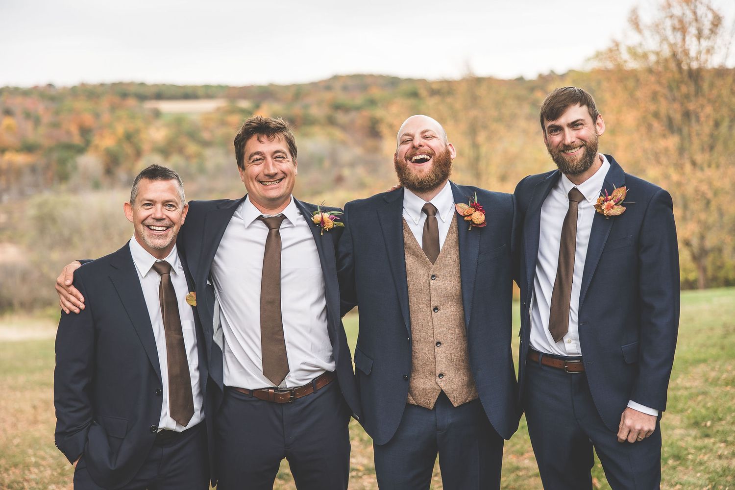 Wedding party men in suits and suspenders smile together against fall landscape.
