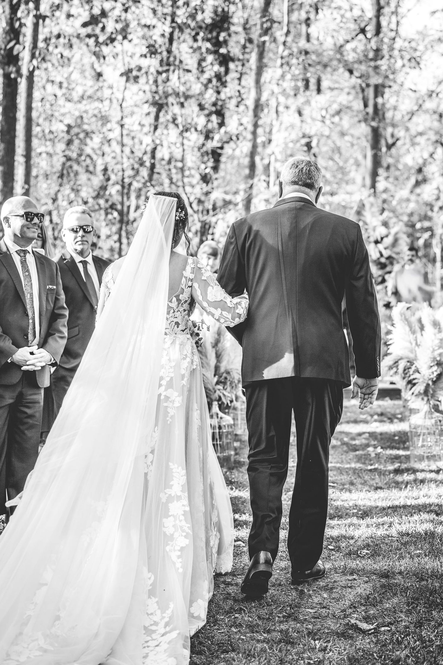 Black and white photo of wedding guests standing in a wooded outdoor ceremony space.