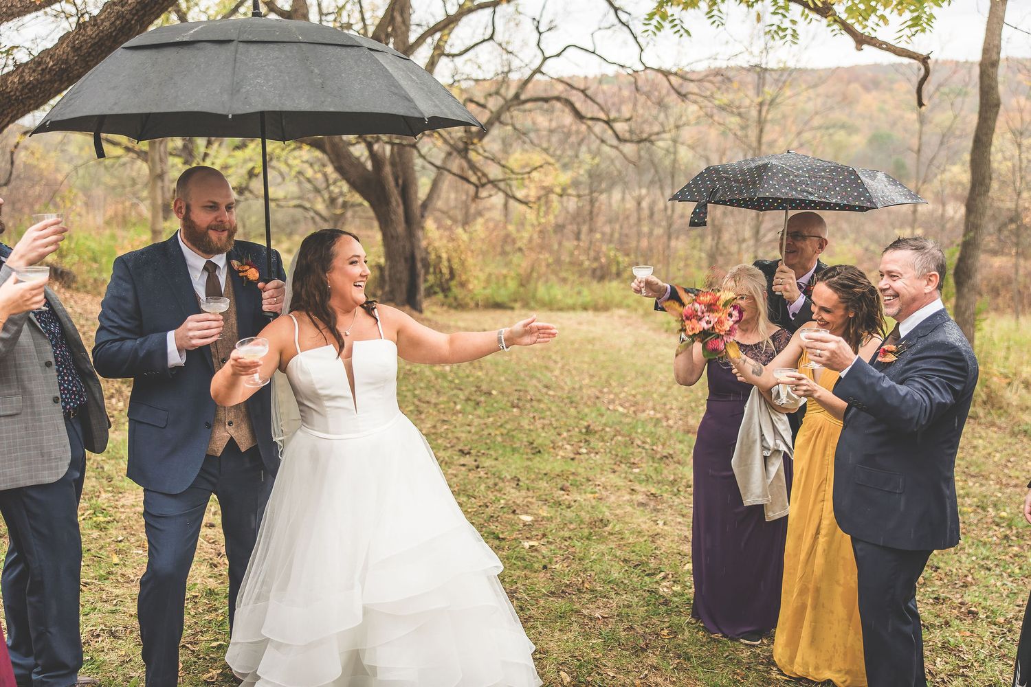Wedding couple shares an intimate moment under umbrellas in a fall forest setting with wedding party in background.