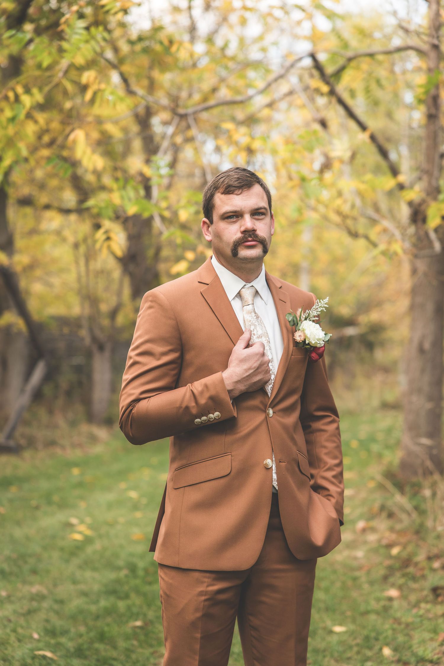 A groom in a tan suit with a boutonniere poses in an autumn forest setting.