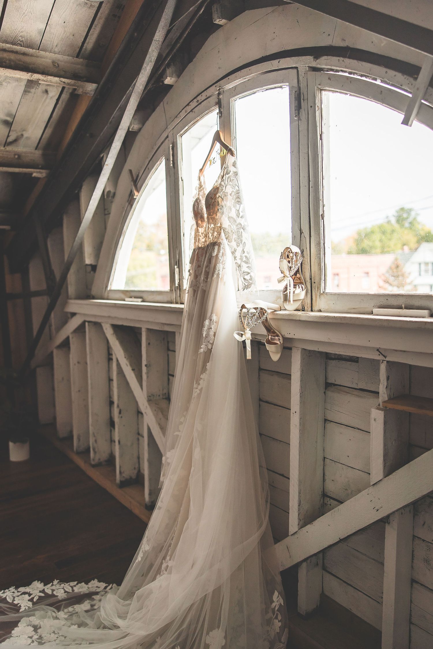 Wedding dress hanging in rustic white arched window frame with natural light streaming in.