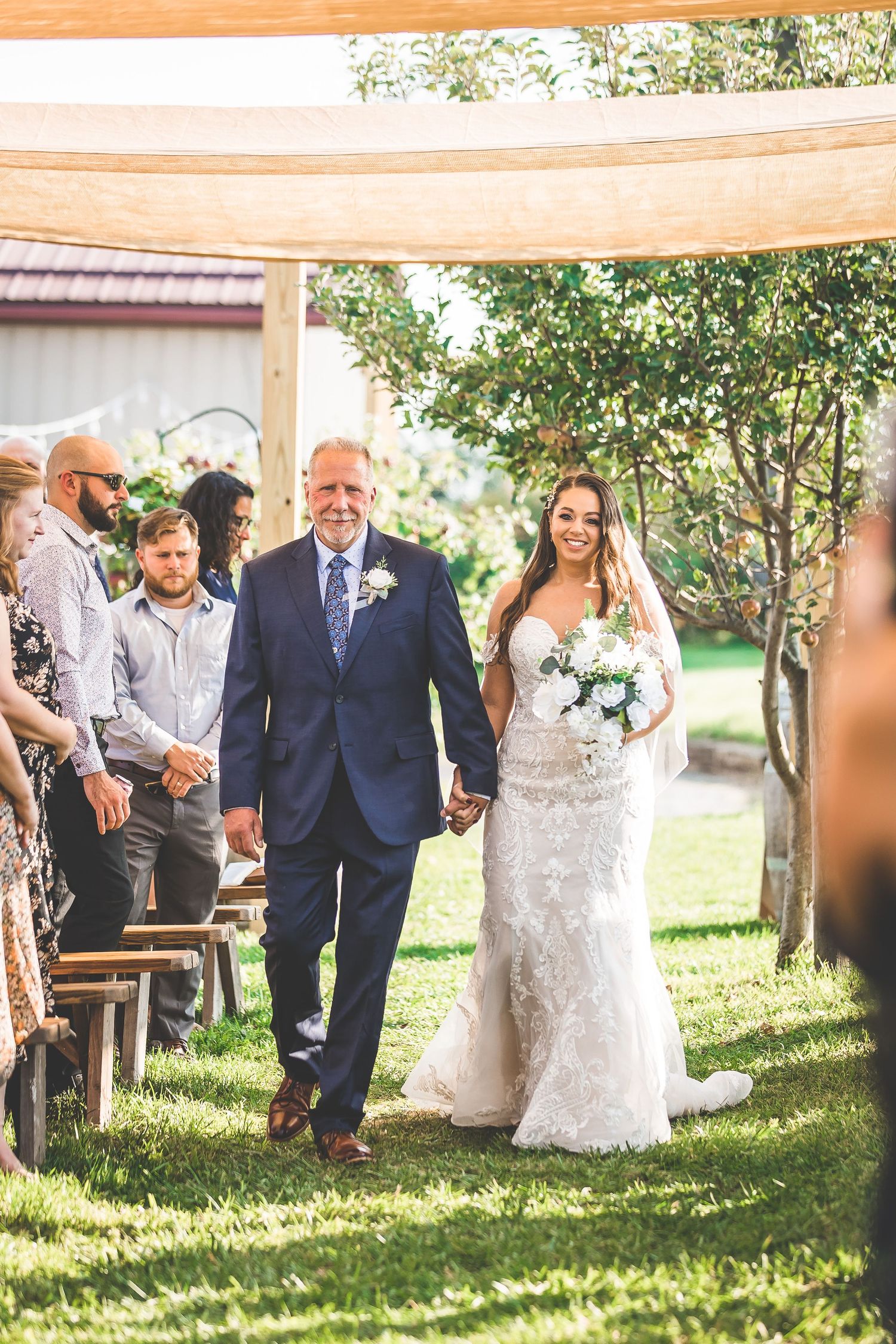 Wedding ceremony guests seated under a white canopy tent in an outdoor garden setting.