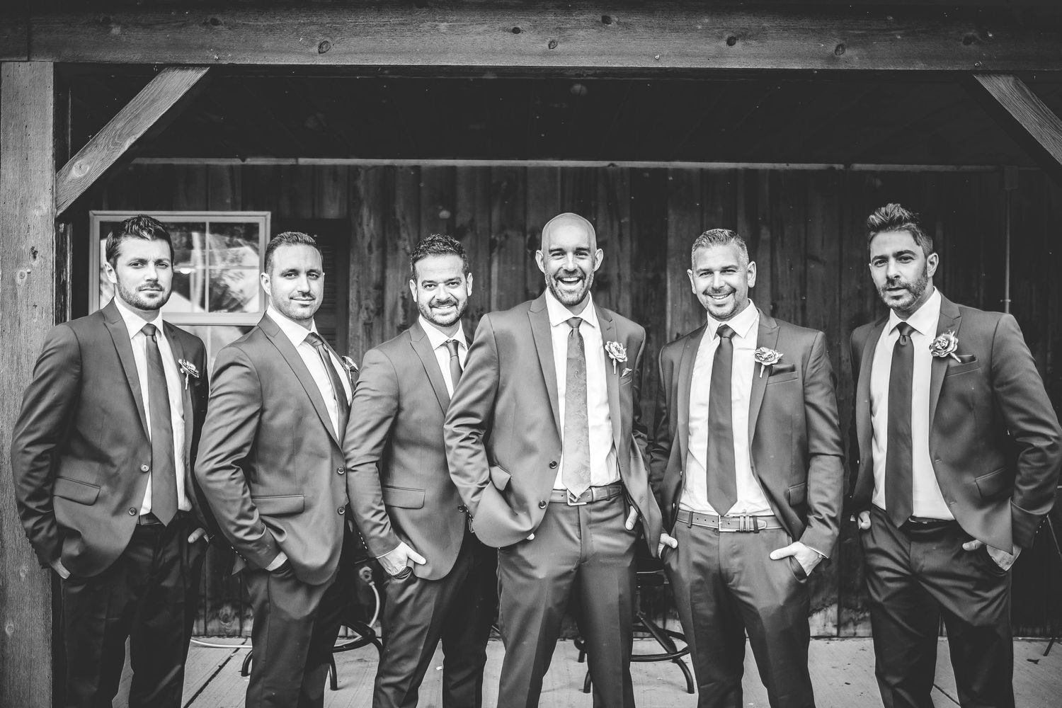 Black and white photo of groomsmen in suits posing against rustic wooden backdrop.