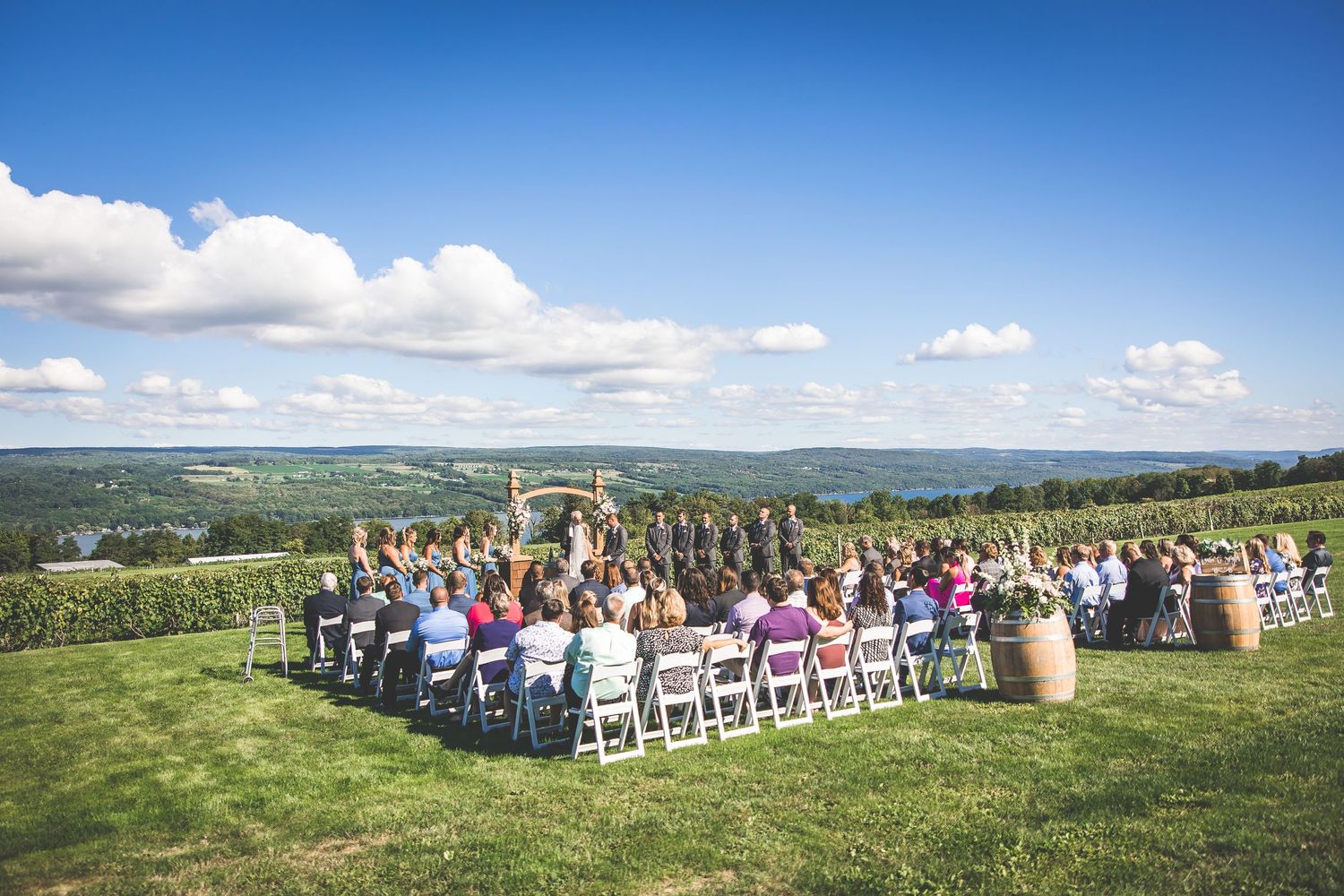 Wide angle view of wedding guests seated in white chairs on a scenic hilltop overlooking valleys.