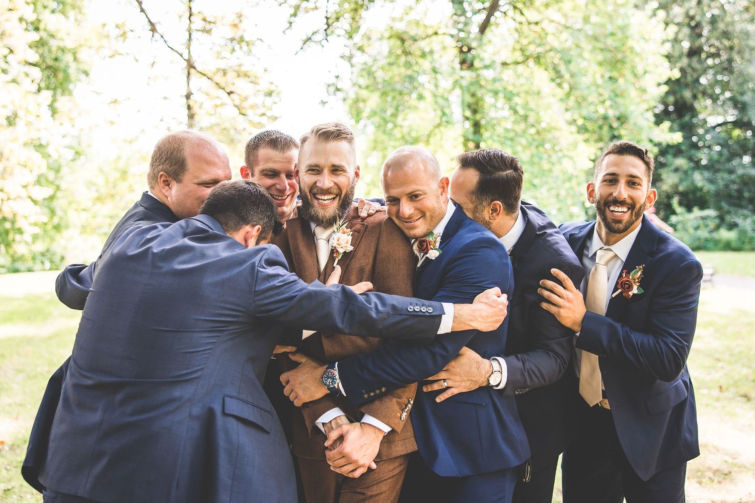 Groomsmen share a joyful group hug in navy suits at an outdoor wedding celebration.