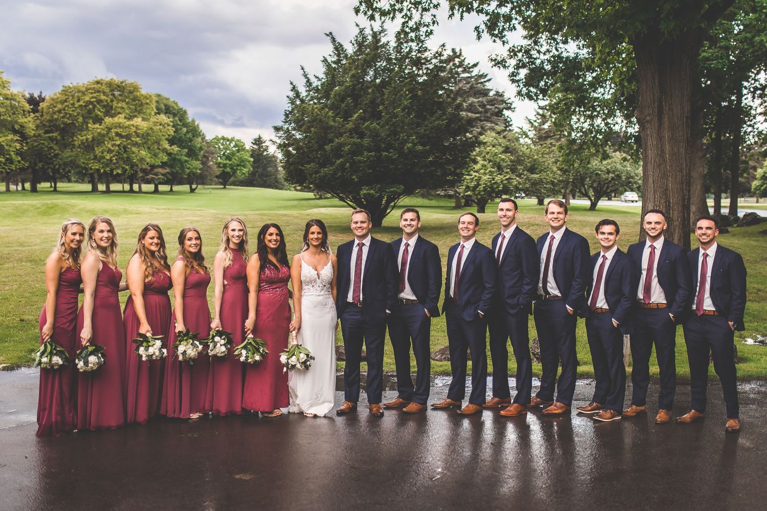 Large wedding party in burgundy dresses and navy suits poses outdoors.