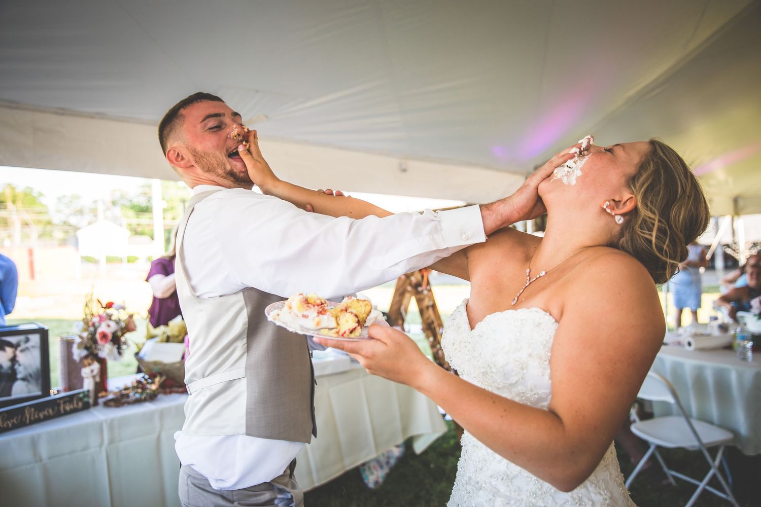 Playful cake-feeding moment between newlyweds at outdoor wedding reception.