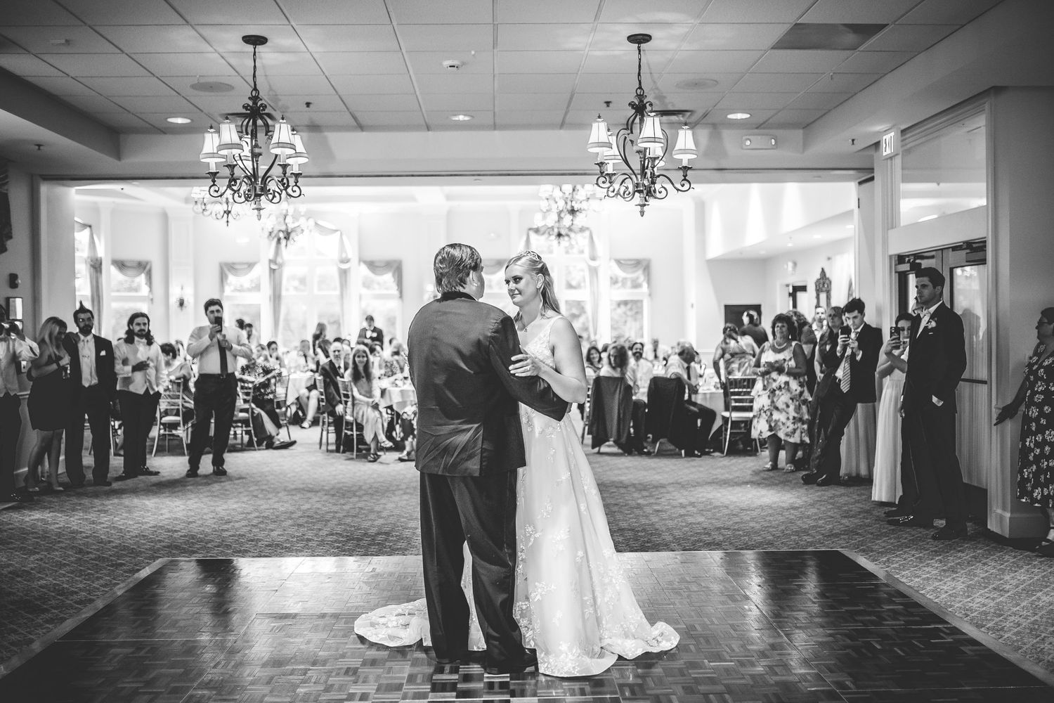 Black and white wide shot of a wedding reception first dance with guests looking on.