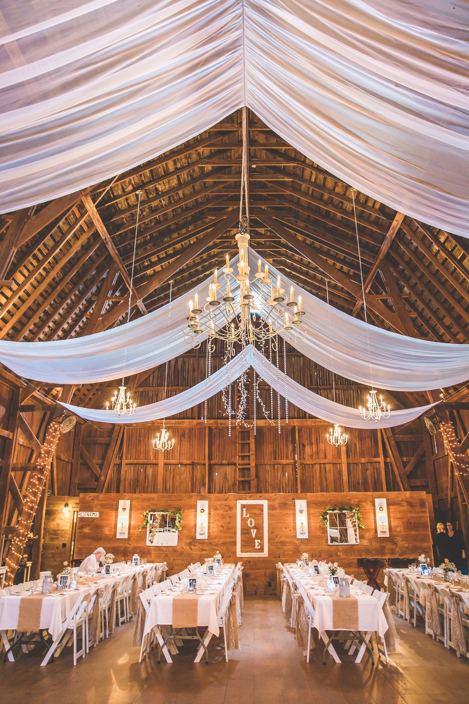 Rustic barn wedding reception venue with elegant white draping and crystal chandeliers.