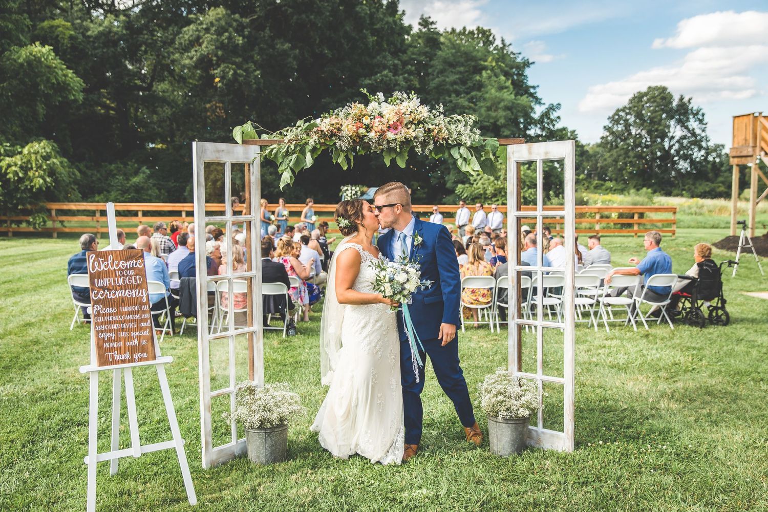 Outdoor wedding ceremony setup with white chairs and wooden arch on a grassy lawn.