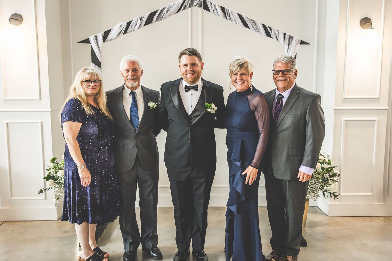 Wedding party members gather for formal group photos in an elegant white room with decorative molding.