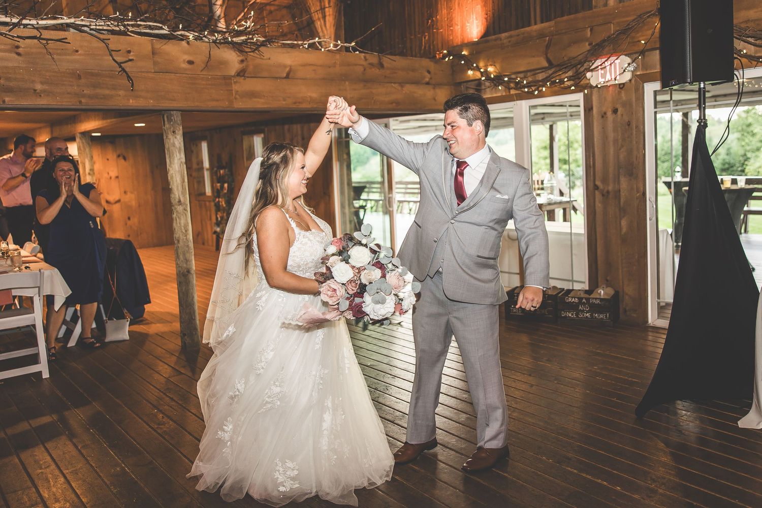 Wedding guests watch as couple dances in rustic barn reception with string lights and wooden details.