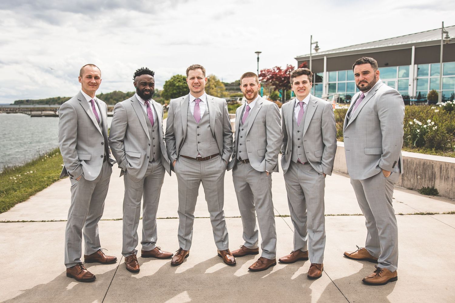 Groomsmen in light gray suits pose together on waterfront walkway.