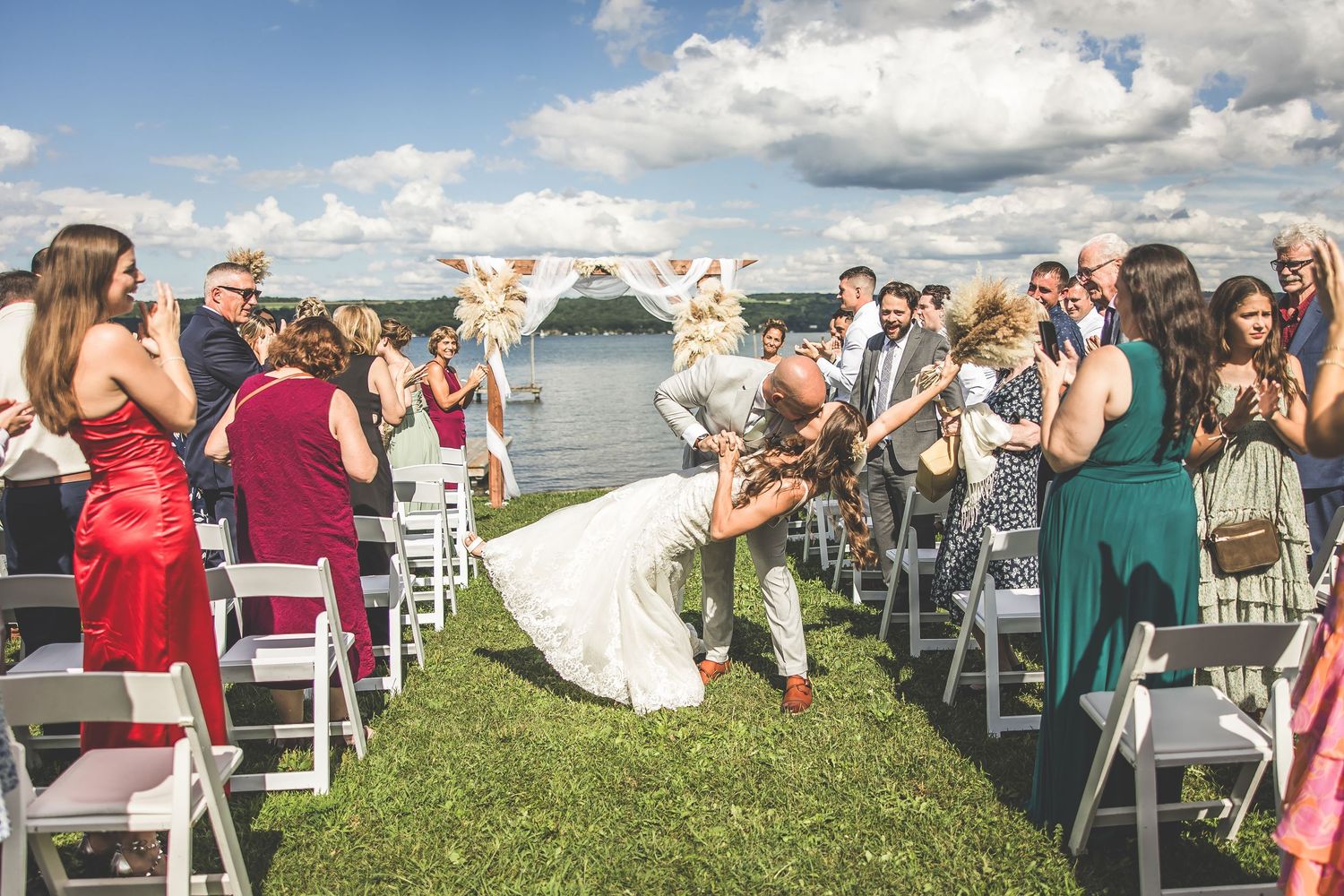 Wedding guests seated in white chairs by the water during an outdoor lakeside ceremony.