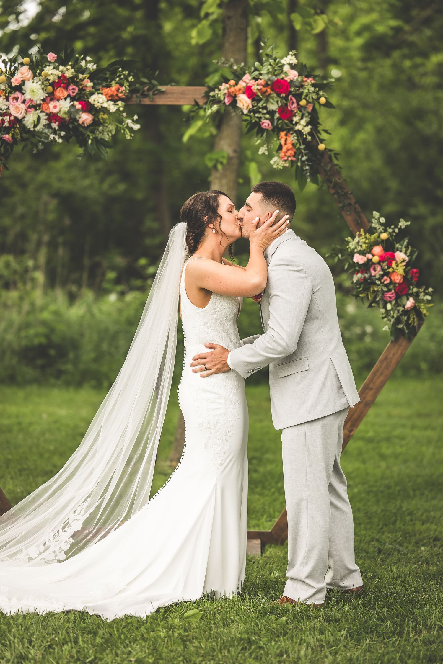 Newlyweds share first kiss under floral arch during outdoor garden wedding ceremony.