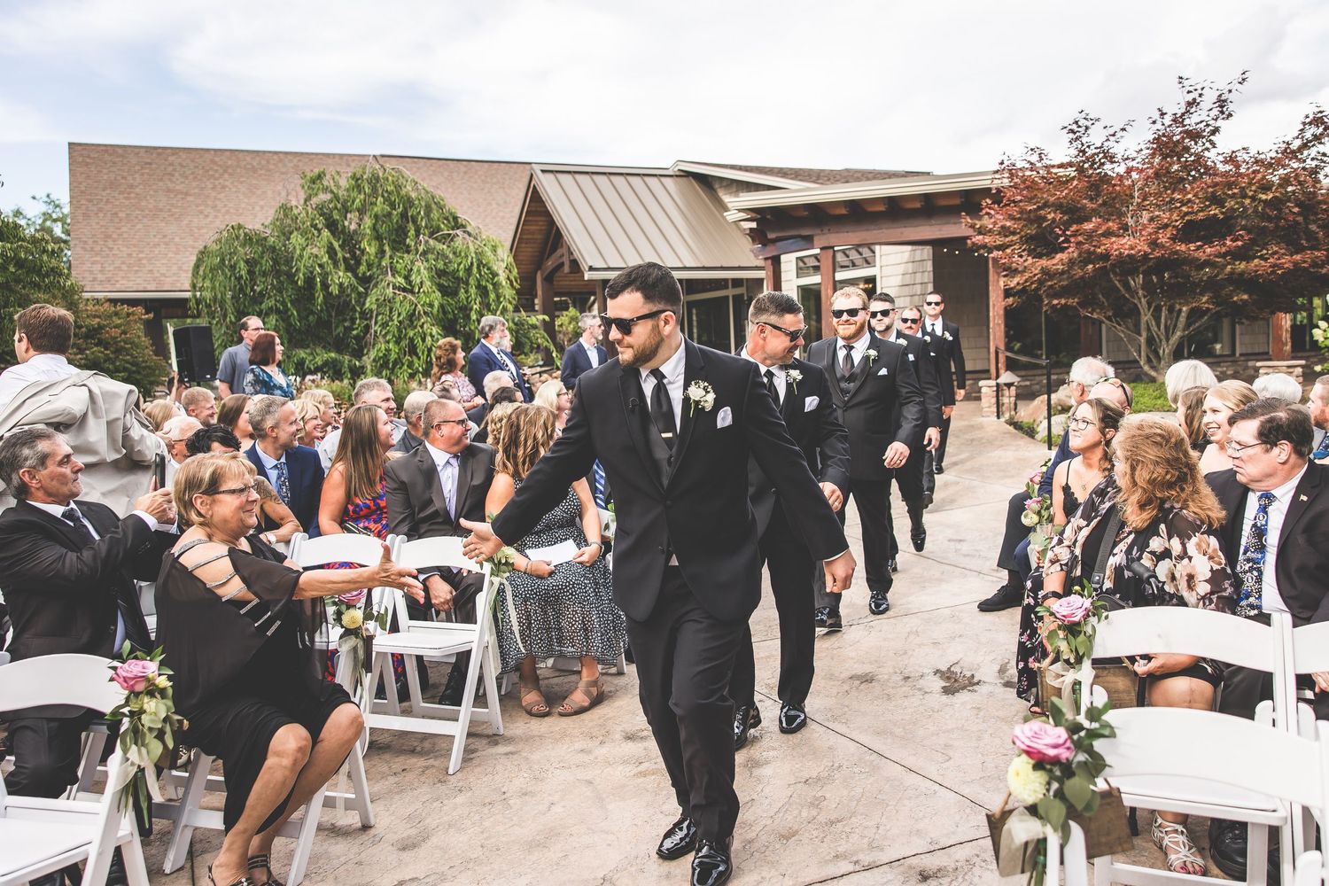 Outdoor wedding ceremony with guests seated in white chairs under natural lighting.