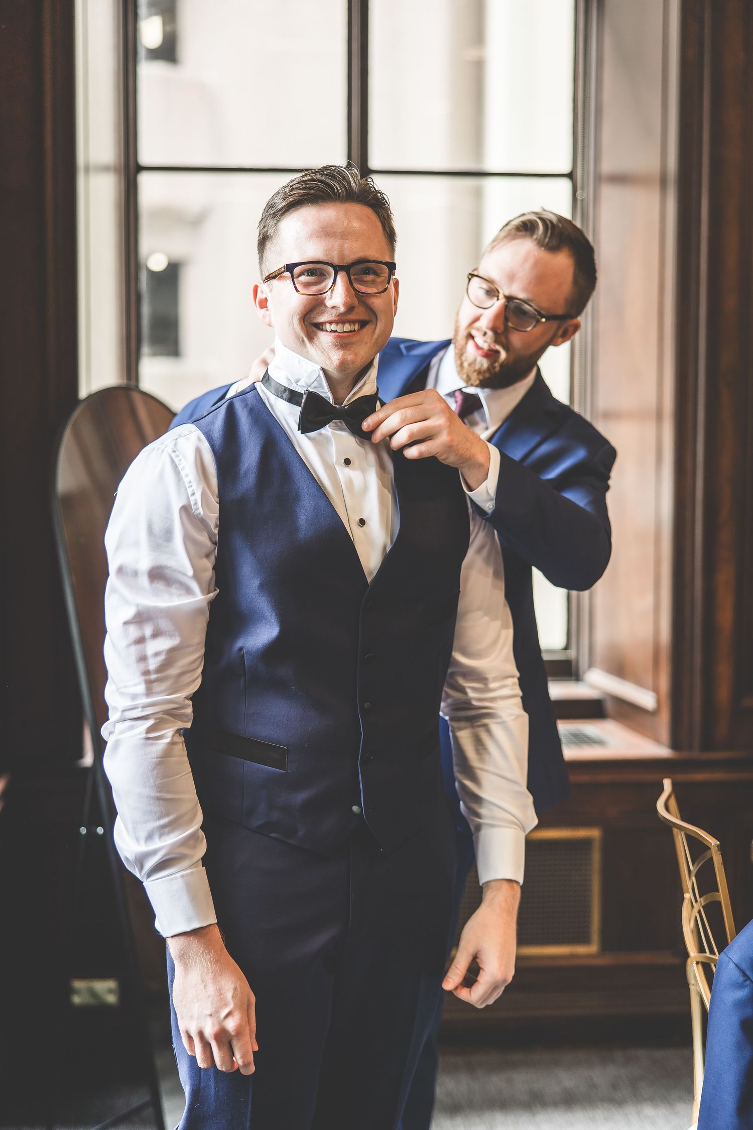 Two people in formal attire share a lighthearted moment while adjusting bowties.