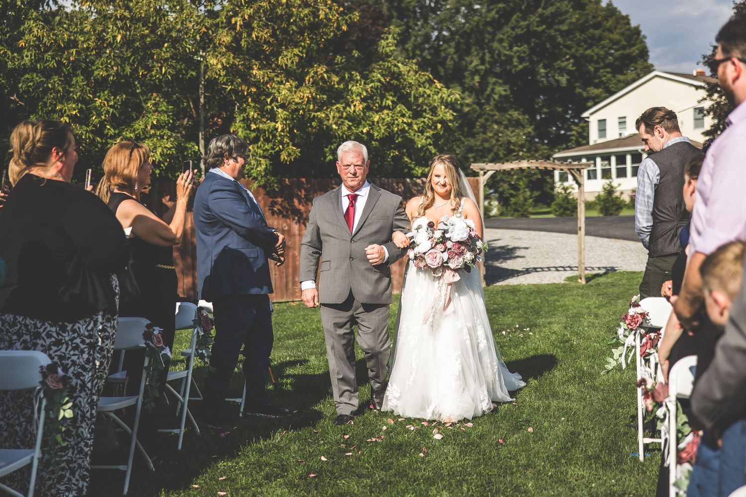Outdoor wedding ceremony processional in front of rustic barn with guests seated on lawn.