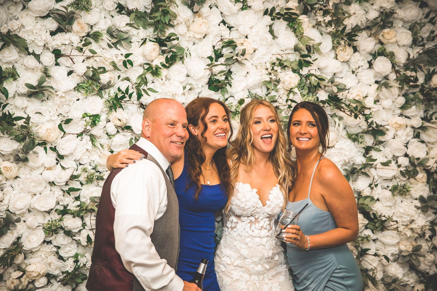 A group poses together against a white floral backdrop at a wedding celebration.