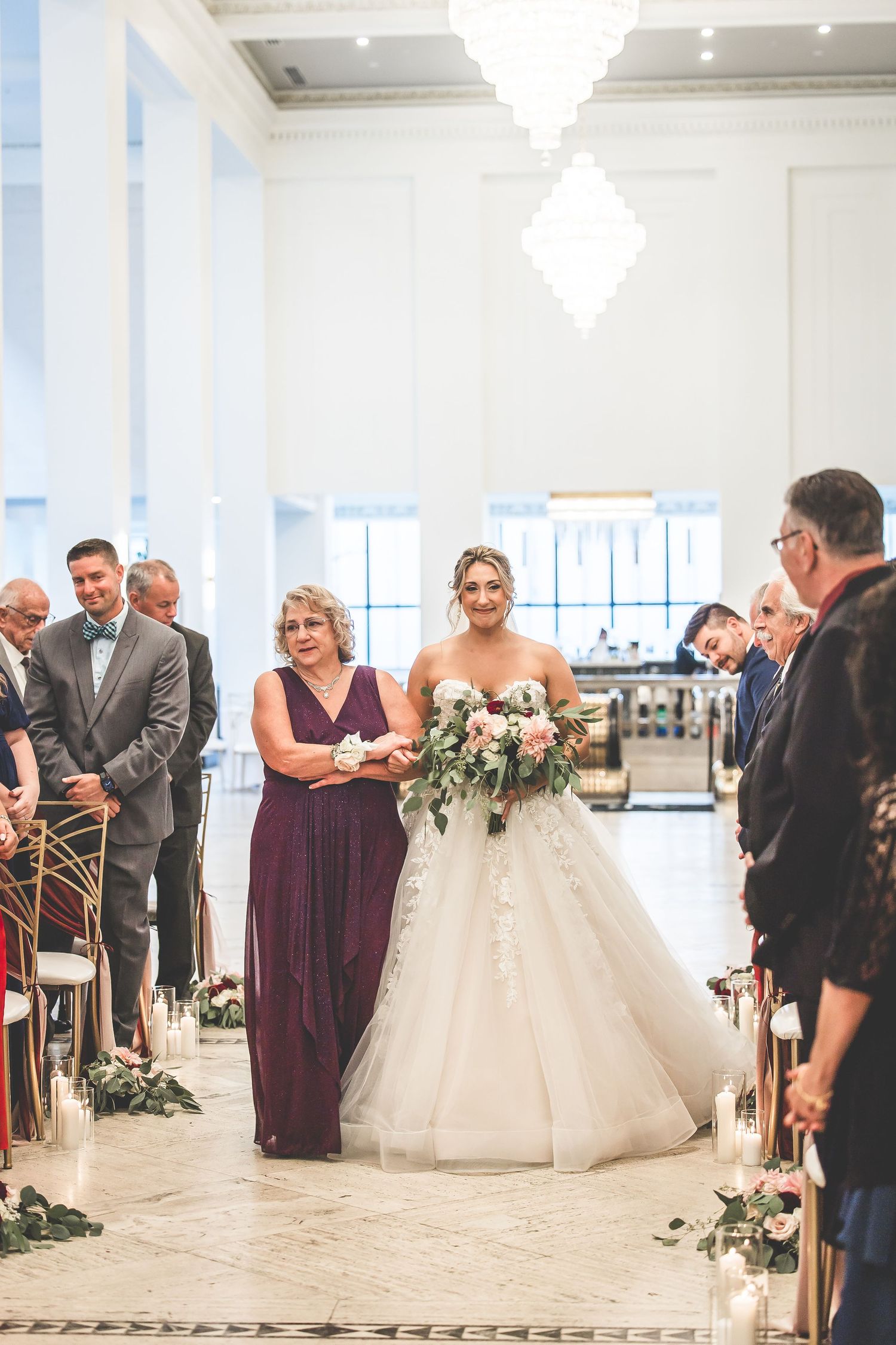 Elegant wedding ceremony setup with guests in a bright white venue featuring chandeliers and floral decorations.