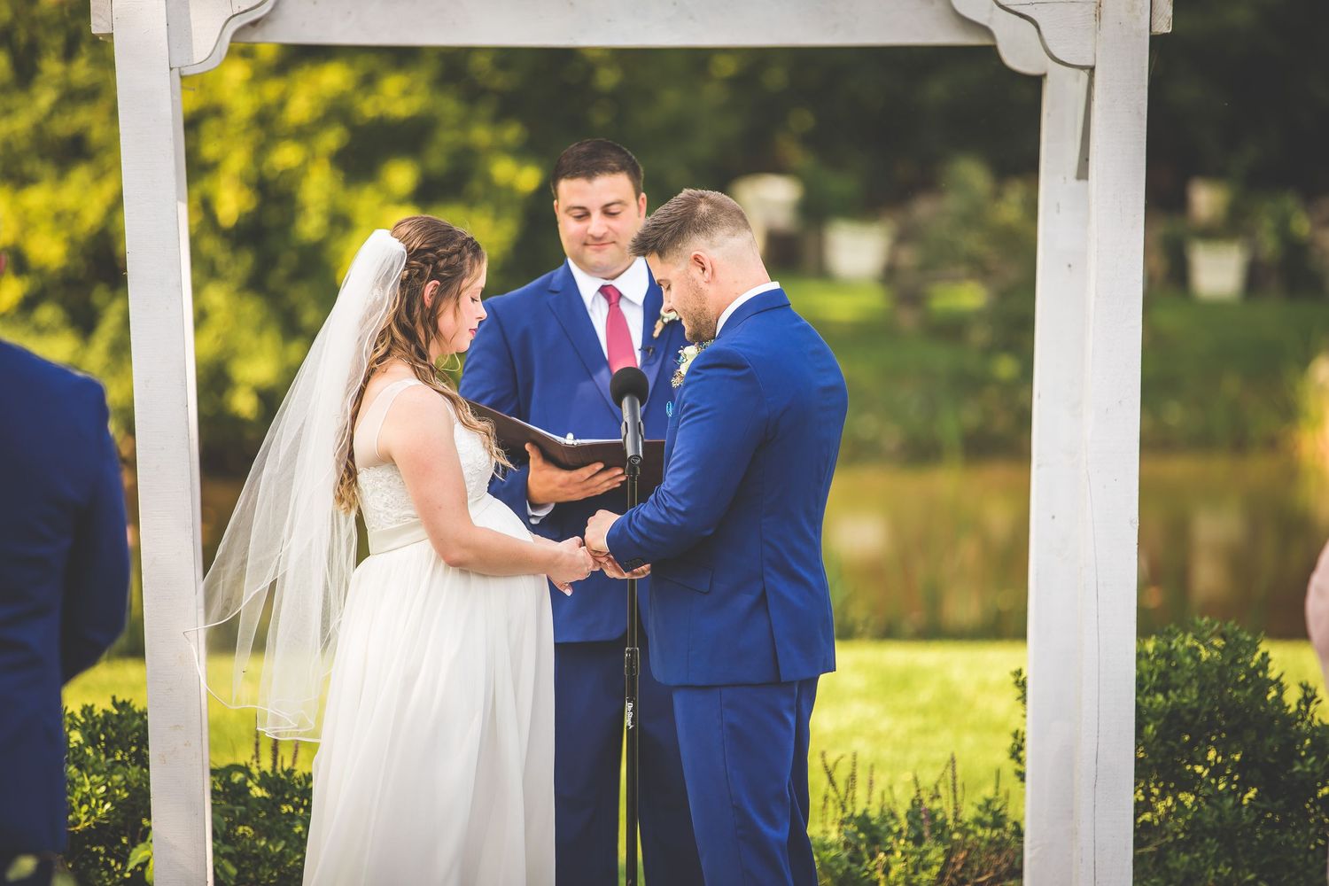 Wedding ceremony taking place under a white pergola with summer foliage in the background.