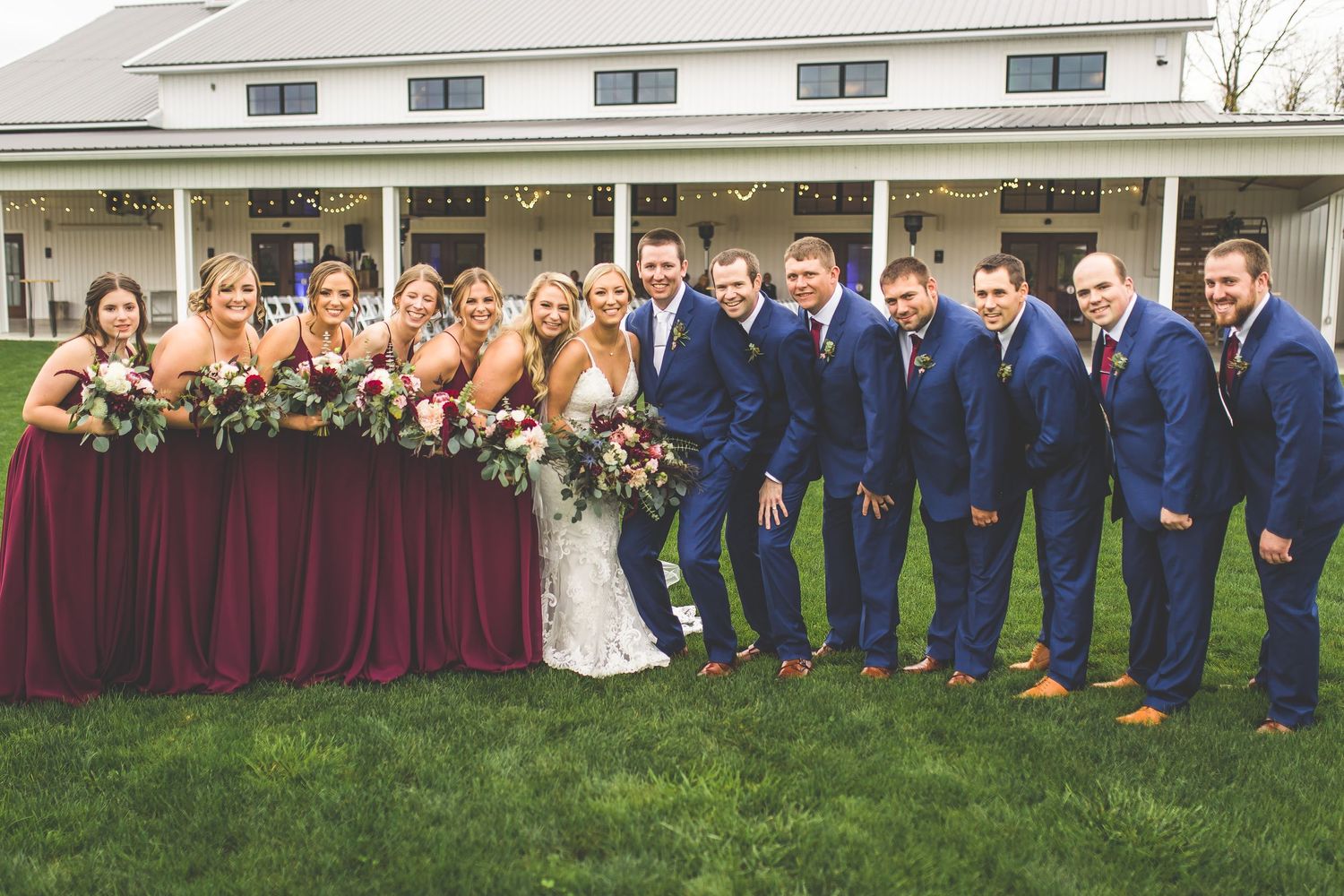 Wedding party poses for group photos in front of a white building with burgundy bridesmaids dresses.